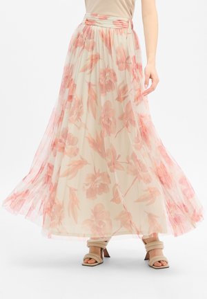 Vloeiende crèmekleurige rok met roze bloemenpatroon, gedragen met beige sandaaltjes met hak en bandjes en een beige top die in de rok is gestopt.