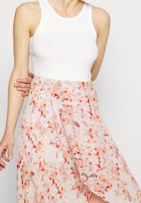 Un débardeur blanc associé à une jupe fluide à motifs de couleur rose clair avec des imprimés floraux rouges, orange et jaunes, présentant un ourlet asymétrique.