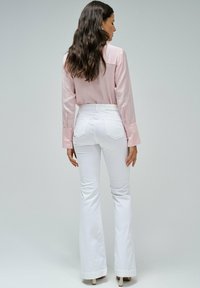 Camisa de manga larga rosa claro con puños amplios, combinada con jeans blancos de tiro alto y acampanados. Textura suave, silueta ajustada y bolsillos decorativos.