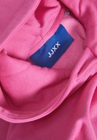 Sweat à capuche rose vif avec étiquette bleue visible à l’intérieur indiquant le nom de la marque JUX.