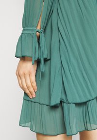 Turkusowa, plisowana sukienka z falbaniastymi rękawami i wiązaniami. Miękka, teksturowana tkanina z warstwowymi falbanami u dołu.