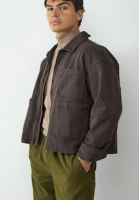 Brązowa bawełniana kurtka z kołnierzykiem, dwoma przednimi kieszeniami i luźnym krojem. Nosi ją się na beżowy sweter z golfem w prążki i zielone spodnie.