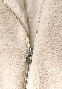 Cremfarbener Fleece-Jacke mit strukturierten Oberfläche, ausgestattet mit einem silbernen Reißverschluss an der Front, der teilweise geöffnet ist und ein beiges Futter zeigt.