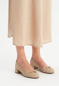 Beige nubuck kengät, joissa on matalat korot, pyöristetyt varpaat ja koristeelliset nauhat etuosassa. Yksinkertainen muotoilu, sileä pinta ja tukeva muoto.