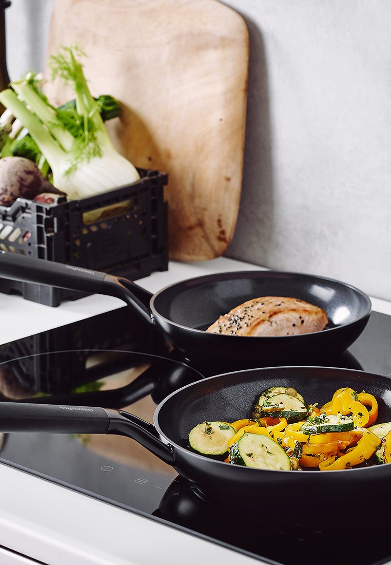 Zwei schwarze Bratpfannen auf dem Herd; eine enthält sautierte gelbe Paprika und Zucchini, die andere hält ein Stück angebratenes Fleisch.