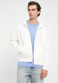 Λευκή φούτερ με φερμουάρ και κουκούλα με κορδόνι, διαθέτει μπροστινές τσέπες και μια διακριτική λεπτομέρεια λογοτύπου. Φοριέται πάνω από ένα ανοιχτό μπλε t-shirt και τζιν παντελόνι.