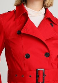 Červený trenčkot s dvouřadým designem, velkými černými knoflíky, detailem opasku a výrazným límcem z texturované látky.