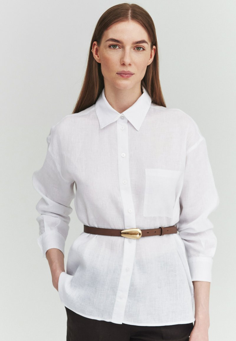 Femme portant une chemise blanche boutonnée, une ceinture marron et un pantalon foncé, debout devant un fond clair uni.