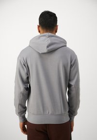 Grå hettegenser med kengurulomma, snorer til hetten og ribbede mansjetter. Stoffet er glatt med en subtil tekstur.