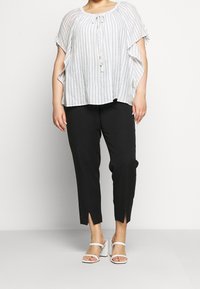 Blusa holgada de rayas blancas y grises con cuello redondo y cierre con cordón, combinada con pantalones negros ajustados con aberturas laterales.