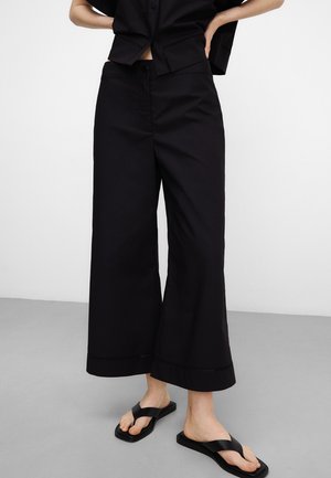 Personne portant un pantalon noir large et court, une chemise noire à manches courtes et des sandales noires, debout sur un sol gris devant un mur blanc.