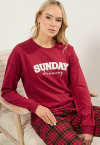 Sudadera roja de manga larga con "SUNDAY" y "dreaming" en texto crema, combinada con pantalones de pijama a cuadros en rojo, negro y amarillo.