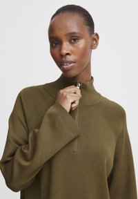 Persona con capelli corti che sta aprendo il colletto di un maglione a maniche lunghe verde oliva, mostrando più orecchini su un orecchio su uno sfondo semplice.