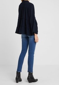 Blouse bleu marine à volants avec de larges manches longues, portée avec un jean foncé ajusté et des bottines noires à cheville avec un ourlet brut.