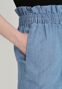 Ljusblå denimshorts med rynkad resår i midjan, med sidofickor och slät textur. Närbild på hand i ficka.