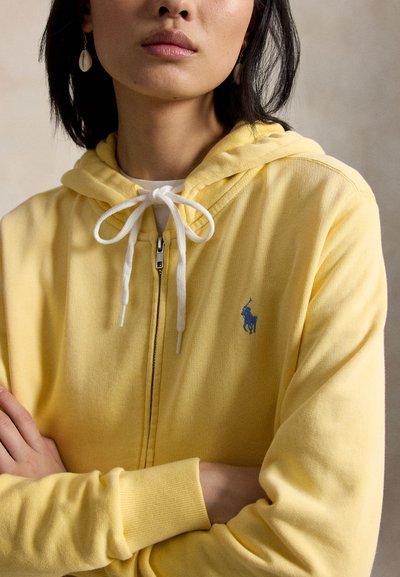 Femme portant un sweat à capuche jaune zippé avec des cordons blancs et un logo Polo Ralph Lauren bleu, bras croisés, portant des boucles d'oreilles en forme de coquillage.
