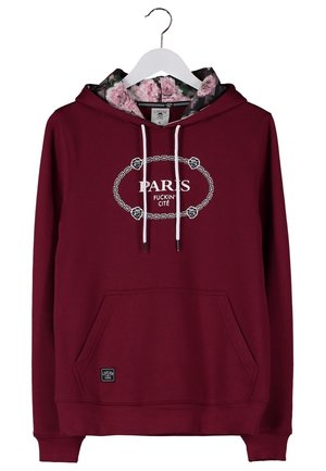 Mann trägt einen dunkelroten Hoodie mit dem Schriftzug „Paris“, blaue Jeans, schwarze Sneakers und hält eine graue Sporttasche an den Griffen.