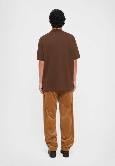 Camisa polo castanha feita de algodão, combinada com calças de corduroy cor de areia, apresentando uma superfície texturizada e ligeiros detalhes em bordado.