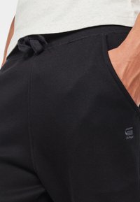 Svarta sweatpants i mjukt material, med dragsko i midjan, sidofickor och en diskret tryckt logga på vänster sida.