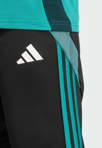 Crne sportske hlače s tirkiznim bočnim akcentima i tri bijele pruge, napravljene od teksturirane tkanine. Istaknuti bijeli logotip.