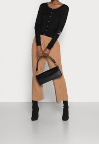 Svart stickad kofta med knappar, ljusbruna vida byxor och en svart läderhandväska med guldfärgade detaljer.