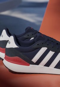 Par de zapatillas Adidas azul marino con rayas blancas, detalles rojos en el talón y suelas blancas colocadas sobre una superficie naranja.