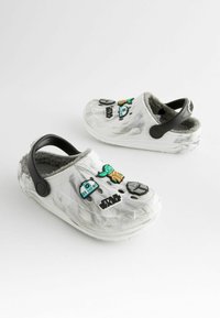 Weiße Clogs mit einer strukturierten Oberfläche, ausgestattet mit schwarzen Akzenten und Star-Wars-Themen-Dekorationsanhängern. Mit grauem Fell gefüttert für zusätzlichen Komfort.