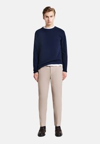 Felpa blu navy con colletto rotondo, indossata sopra una camicia bianca, abbinata a pantaloni beige chiaro e scarpe scure. Design semplice e aderente.