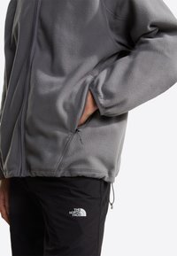 Persoon draagt een grijze fleecejas met hand in de zak en zwarte broek met het logo van The North Face op de dij.