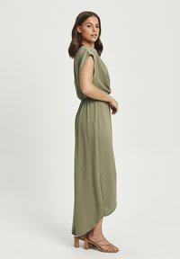 Olive green maxi dress with short sleeves and a draped design. Features a cinched waist and flowing texture. Worn with beige strappy sandals.