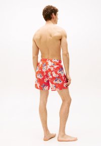 Costum de baie scurt roșu cu motive florale, având un elastic la talie, prezentând modele mari cu flori albe și albastre, văzut din spate pe un tors gol.