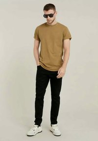 Κοντομάνικη, μουστάρδη μπλούζα, μαύρο στενό παντελόνι, λευκά αθλητικά παπούτσια και σκούρα γυαλιά ηλίου, να στέκονται μπροστά από ένα ουδέτερο φόντο.