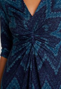 Laivastonsininen ja merensininen mekko V-kaula-aukkolla, kolme neljäsosaa hihoja, rypytetyllä yläosalla ja teksturoidulla, kimaltelevalla kankaalla geometrisessa kuviossa.