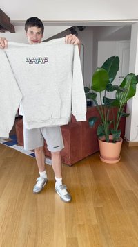 Szürke pulóver, multicolor szöveges mintával, szürke rövidnadrággal párosítva. A modell egy fából készült padlón áll, beltéri növények és egy kanapé mellett.