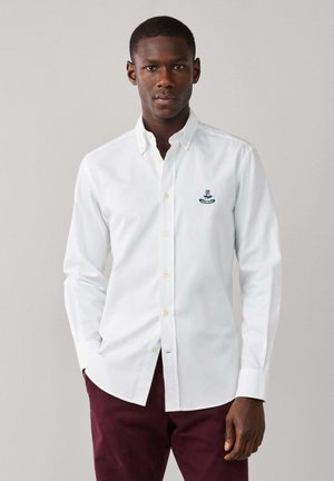 Man wearing a white button-up shirt with a small embroidered crest on the chest and dark red pants standing against a plain background.