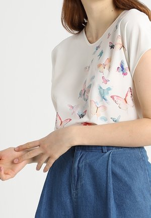 Vrouw draagt een witte blouse met kleurrijke vlinderprints en een hoogwaterbroek in blauw, ze past haar linkerpols aan met haar rechterhand.