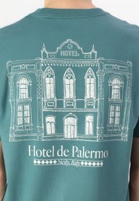 Camiseta de manga corta en color teal con una ilustración en contorno blanco de la fachada de un hotel con ventanas arqueadas y acentos decorativos, etiquetada "Hotel de Palermo."