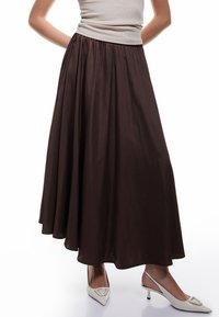 Bruine maxi rok met een geplooide tailleband, gemaakt van een gladde stof. Gecombineerd met witte slingback schoenen met een geometrisch detail.