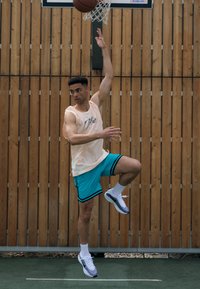 Basketballspieler in beigem ärmellosem Oberteil und türkisfarbenen Shorts, der für den Korb springt, mit weißen und schwarzen Turnschuhen, vor einer Holzspielfläche.