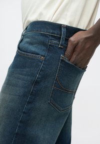 Τζιν παντελόνι σε σκούρο μπλε με φθαρμένο εφέ. Διαθέτει κανονική μέση, πλαϊνές τσέπες και αντίθεση στη ραφή με πορτοκαλί χρώμα.