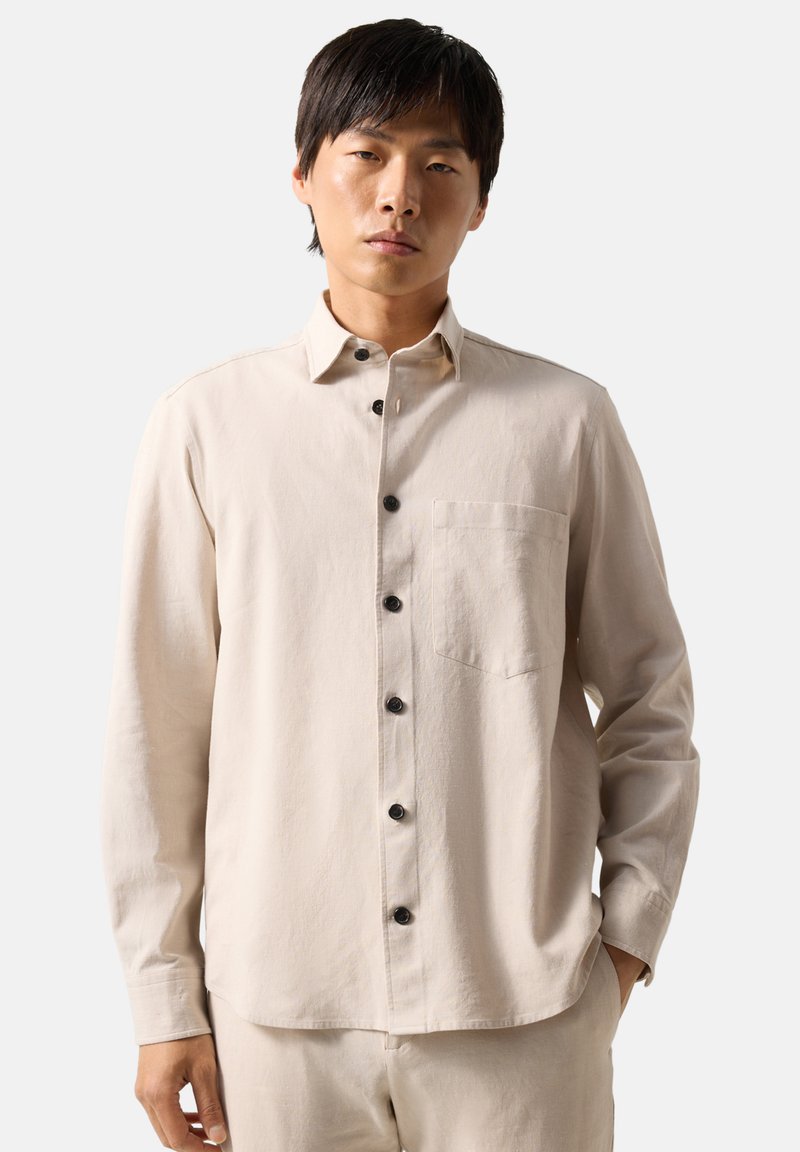 Young man with dark hair wearing a beige long-sleeve button-up shirt with black buttons and matching beige pants.