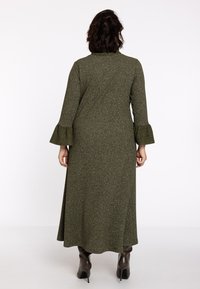 Robe longue olive avec un tissu texturé, des manches évasées et une coupe ample. Associée à des bottes marron jusqu'aux genoux.