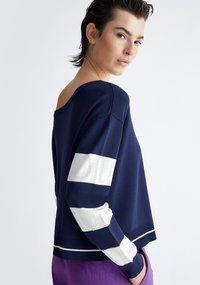 Maglione blu navy con schiena scoperta, maniche a righe bianche con aperture e orlo a coste. Il tessuto appare liscio e morbido.