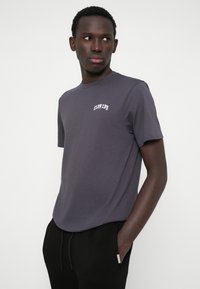 T-shirt cinza de gola redonda feita em algodão, com mangas curtas e um pequeno logo branco bordado no peito. Textura suave.