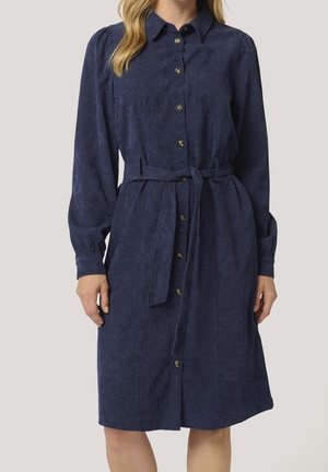 Femme portant une robe chemise bleu marine à manches longues avec une ceinture nouée à la taille, debout devant un fond uni.