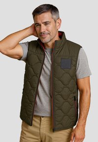 Olivengroene gewatteerde vest met een hoge kraag, voorkant met rits en rode accenten. Draag het over een grijs T-shirt, met twee zijzakken.