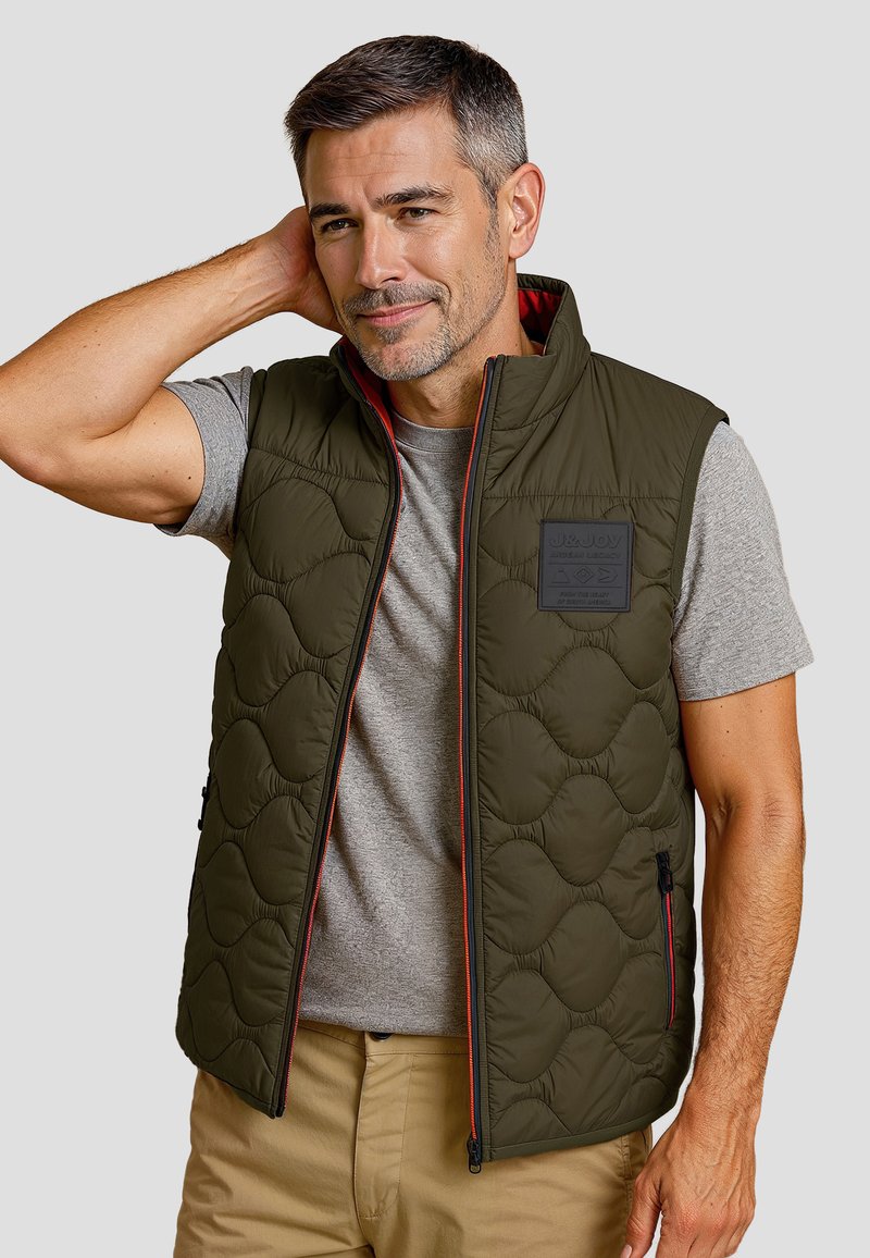 Olivengroene gewatteerde vest met een hoge kraag, voorkant met rits en rode accenten. Draag het over een grijs T-shirt, met twee zijzakken.