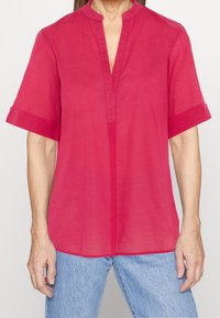 Blusa rosa vivace a maniche corte con colletto alla mandarina e bottoni parziali, realizzata in tessuto leggero, abbinata a jeans blu.