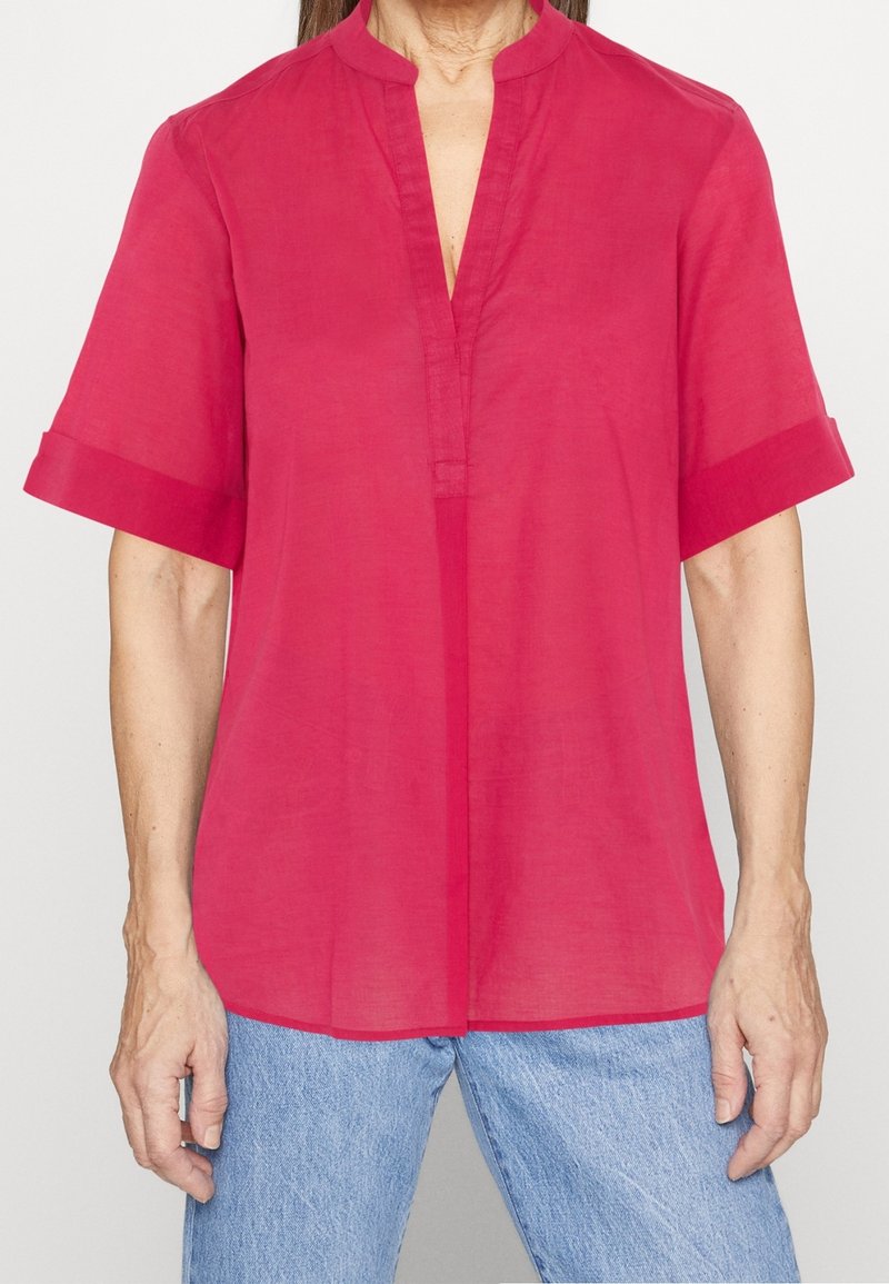 Blusa rosa vivace a maniche corte con colletto alla mandarina e bottoni parziali, realizzata in tessuto leggero, abbinata a jeans blu.