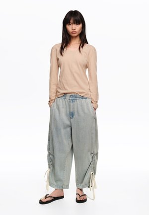 Kobieta w beżowej bluzce z długim rękawem, jasnoniebieskich szerokich spodniach jeansowych o długości do połowy łydki z wiązaniami oraz czarnych sandałach na platformie, stojąca z rękami w kieszeniach.